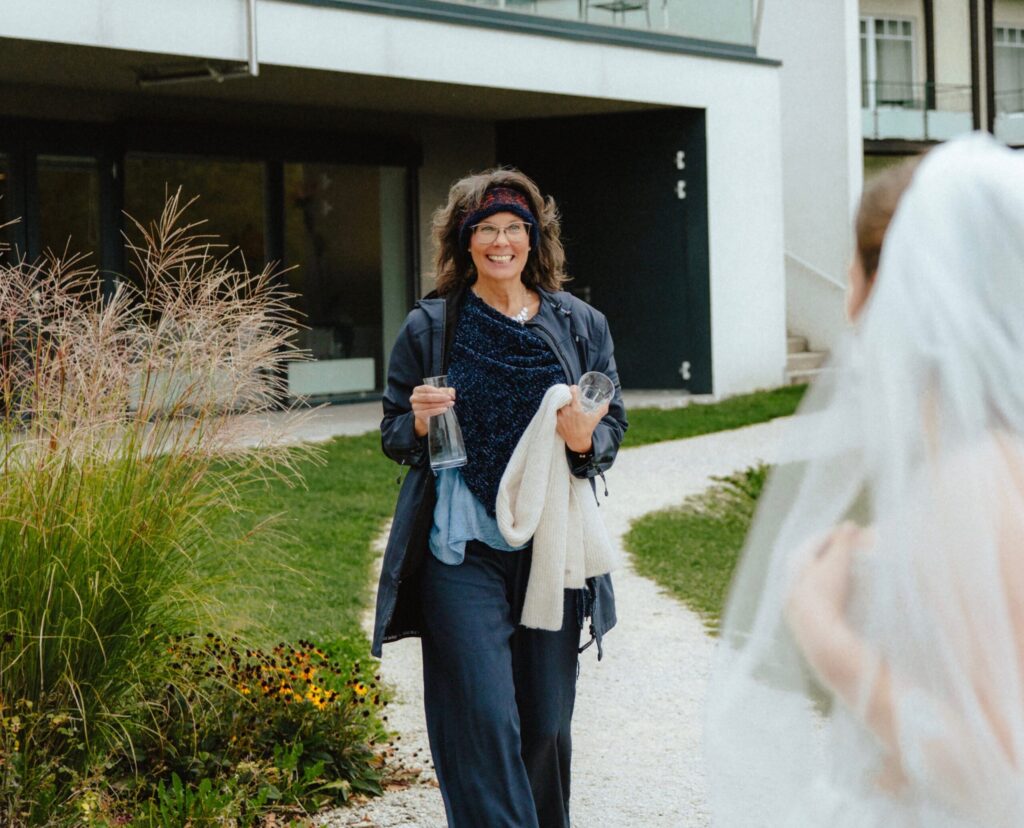 Frau bei einer Hochzeit in Österreich, die als freie Rednerin spricht, mit einem Glas in der Hand, vor modernem Gebäude, umgeben von Natur.