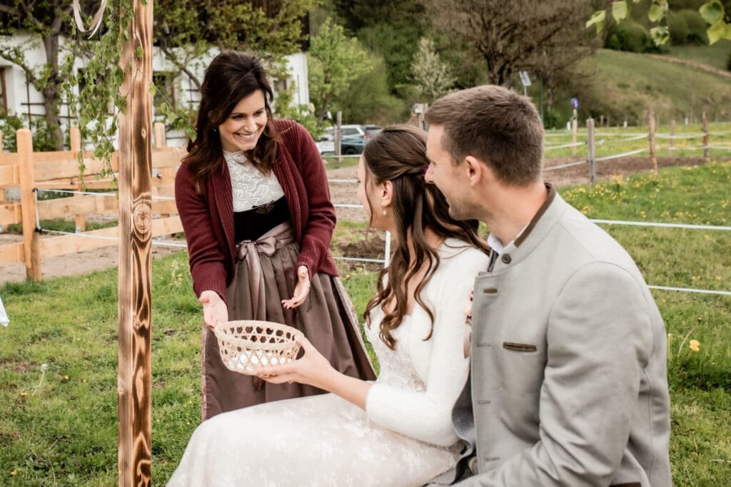 Brautpaar bei einer Freiluft-Hochzeit in Österreich, mit einer Rednerin, die eine Zeremonie im Grünen durchführt, umgeben von Natur und Garten.