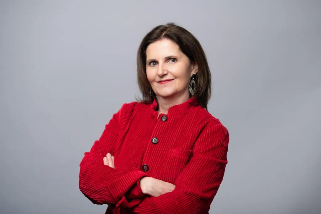 Rednerin Maria Reingruber in rotem Blazer, die selbstbewusst und professionell wirkt, steht vor einem neutralen Hintergrund. Sie symbolisiert Kompetenz und Erfahrung im Bereich öffentliches Reden und Präsentationen in.