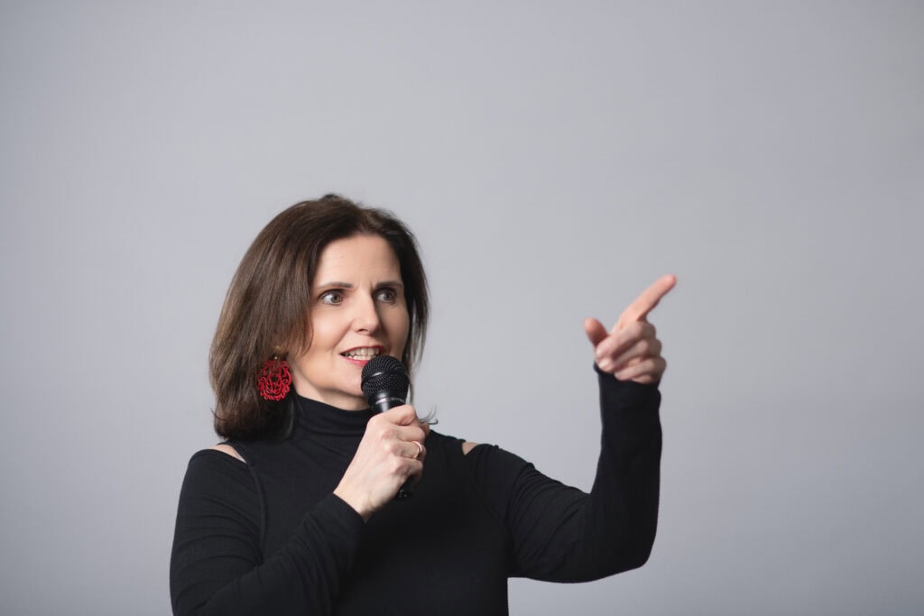 Freie Rednerin Maria Reingruber spricht bei einer Veranstaltung in Österreich, hält ein Mikrofon und zeigt mit der Hand nach oben, um Aufmerksamkeit zu erregen.