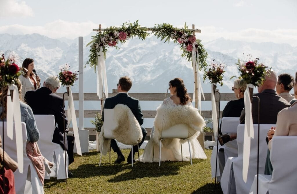 Hochzeit im Freien mit Bergpanorama, Brautpaar und Gästen unter einem Blumenbogen, umgeben von Natur und Bergen, perfekte Kulisse für eine unvergessliche Trauung.