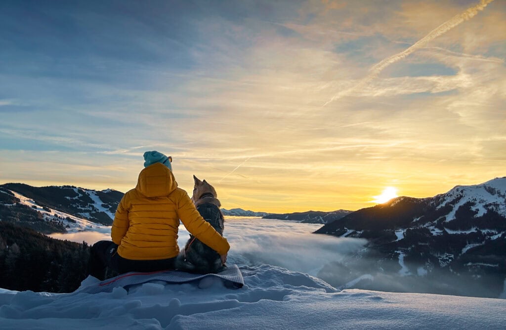 Person und Hund sitzen auf Schneehügel und genießen den Sonnenuntergang in den österreichischen Alpen.