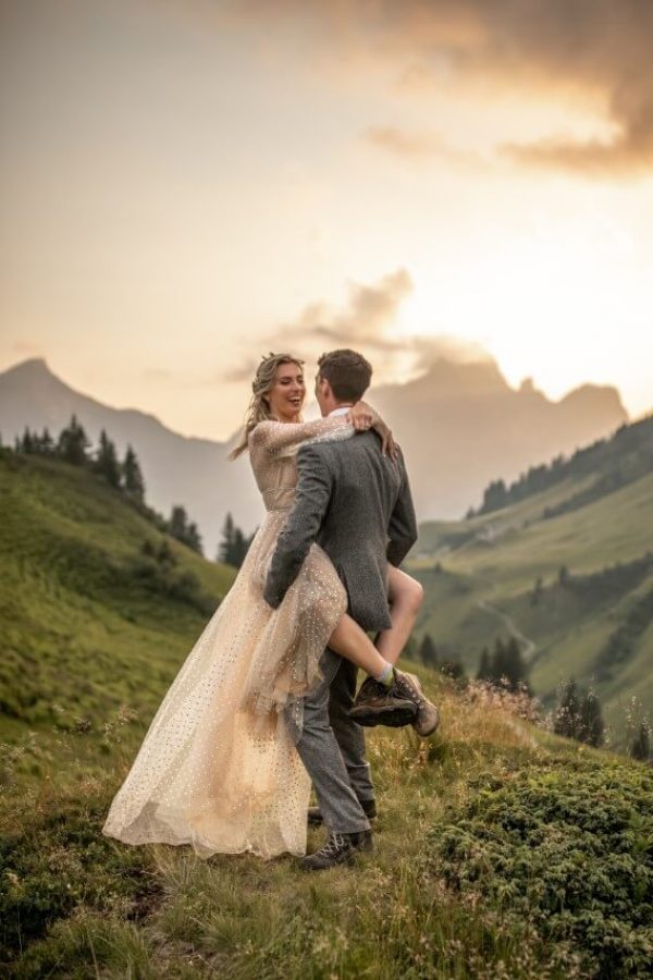 Brautpaar umarmt sich auf einem Berg. Die Braut trägt Wanderschuhe.